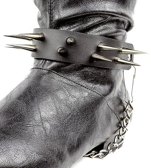 close up shot of black boot displaying black boot strap  that has two rows of multiple silver sharp pointy spike studs and hanging silver chain