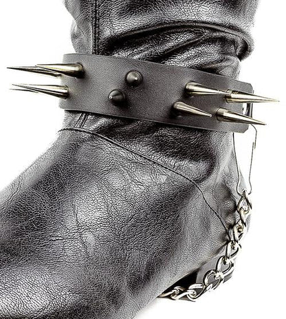 close up shot of black boot displaying black boot strap  that has two rows of multiple silver sharp pointy spike studs and hanging silver chain