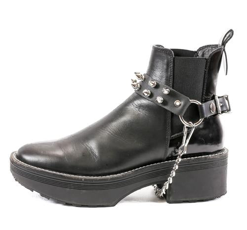 black ankle boot displaying black leather boot strap with two rows of silver tree spikes, single silver o ring and silver hanging chain
