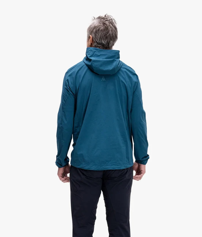 A man viewed from behind wearing a teal blue hooded jacket and dark pants against a plain white studio background.