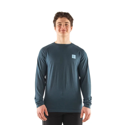 A young man wearing a dark slate blue long-sleeve performance shirt with a small square logo on the left chest, standing against a solid white background.